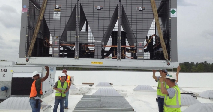 Workers in safety vests carefully position a large air conditioning unit on a rooftop, with cloudy skies in the background.