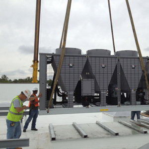 Workers guide a large HVAC unit being lifted onto a roof by a crane, with cloudy skies in the background. Safety gear is visible.